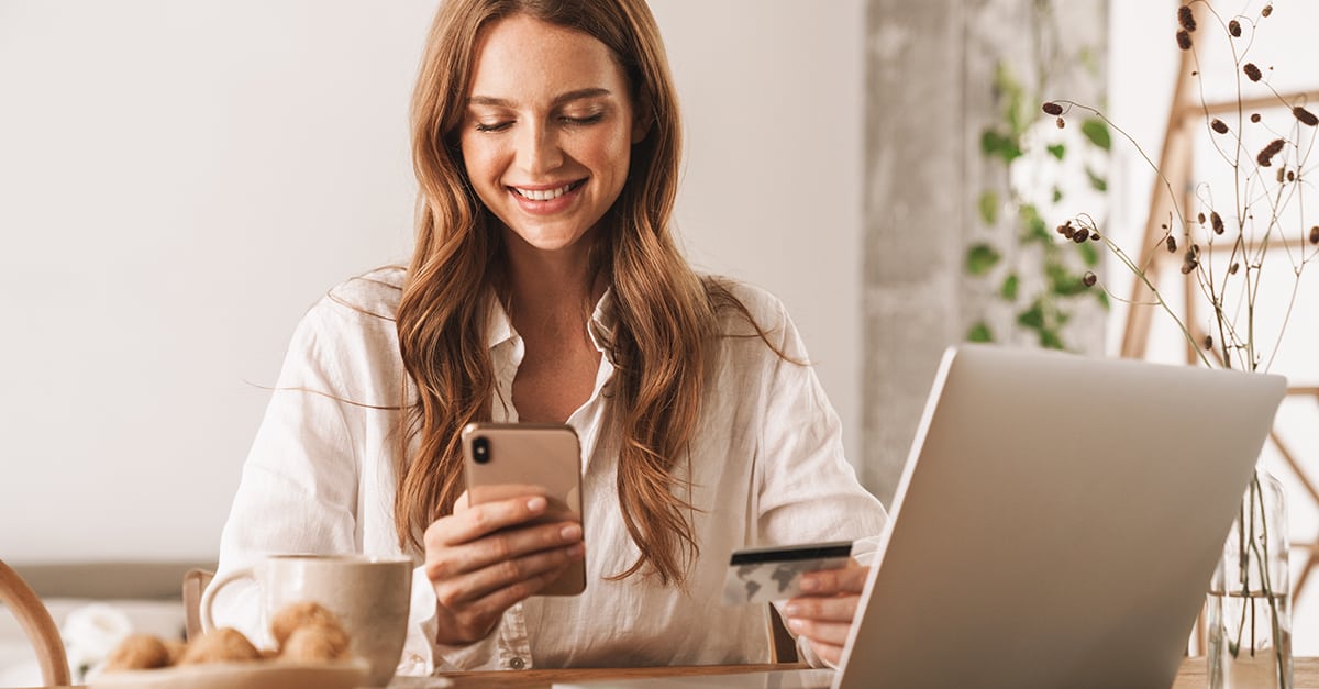 Image of young woman sitting indoors at a table using laptop computer and mobile phone holding credit card.